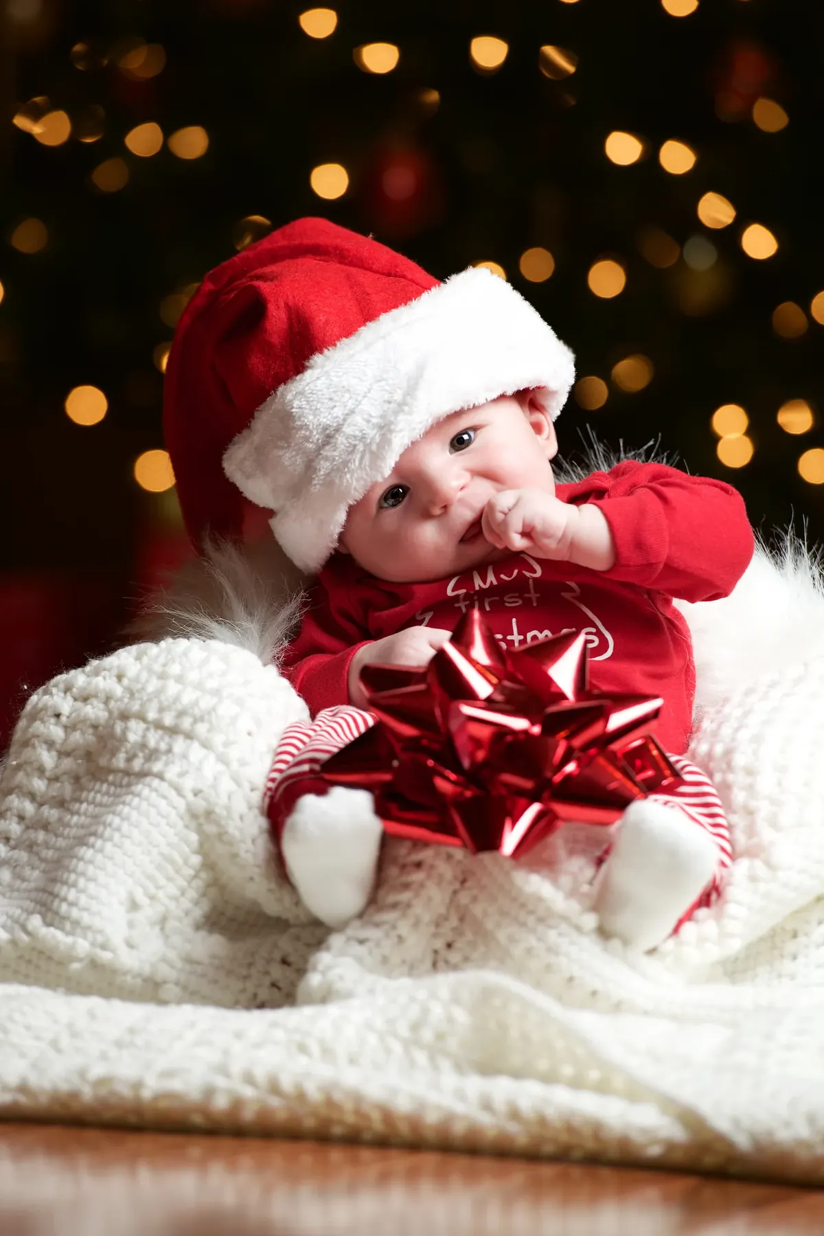 Baby wearing a Santa hat with a red gift bow and warm holiday lights