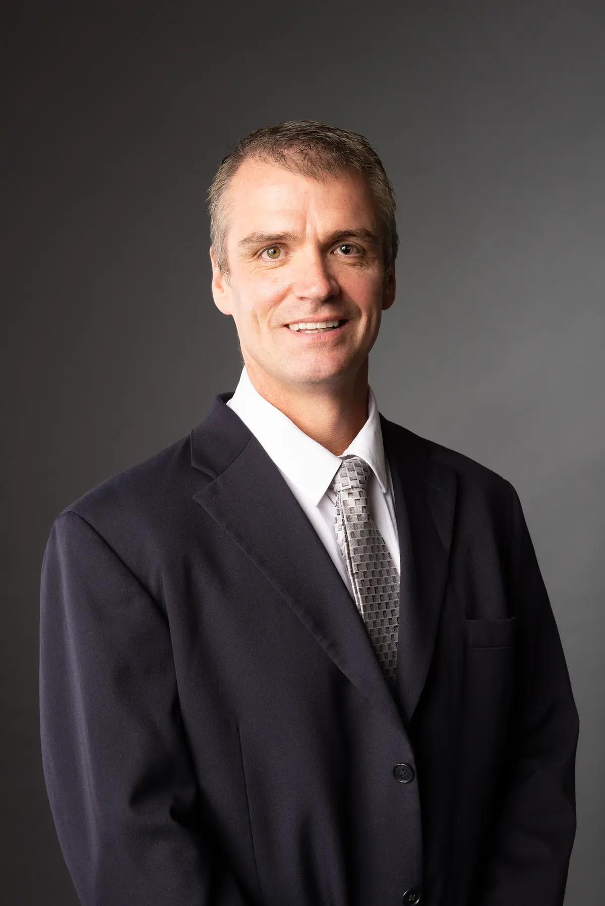 Man in a dark suit and patterned tie against a gray background