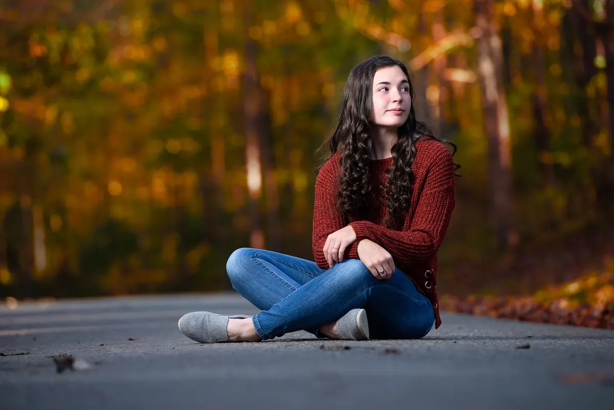 Senior in a burgundy sweater seated on a road with autumn foliage